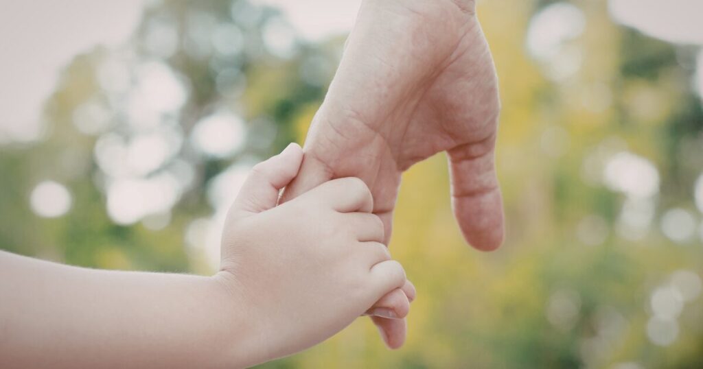 Baby's small hand grips an adult finger, signaling care and bonding. (outdoor background)