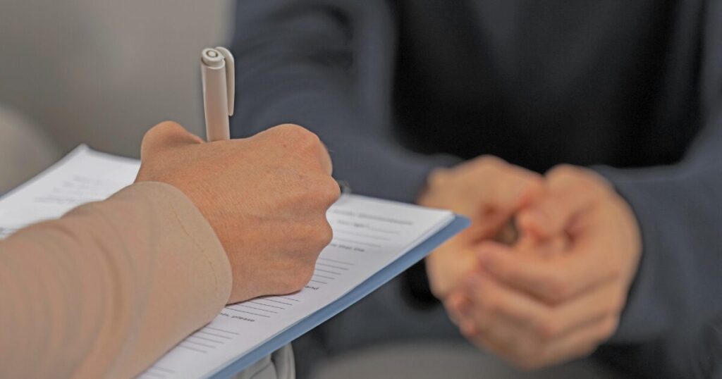 Person signs a document on a clipboard while the other person sits with hands clasped in the background, suggesting an agreement moment away from the camera.