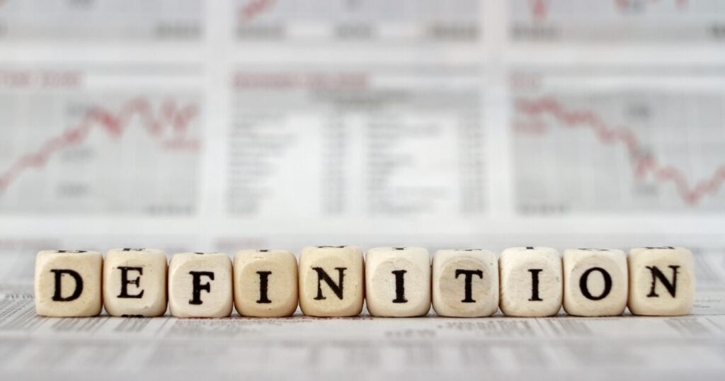 Wooden letter blocks spell'DEFINITION' on a desk with blurred financial charts in the background.