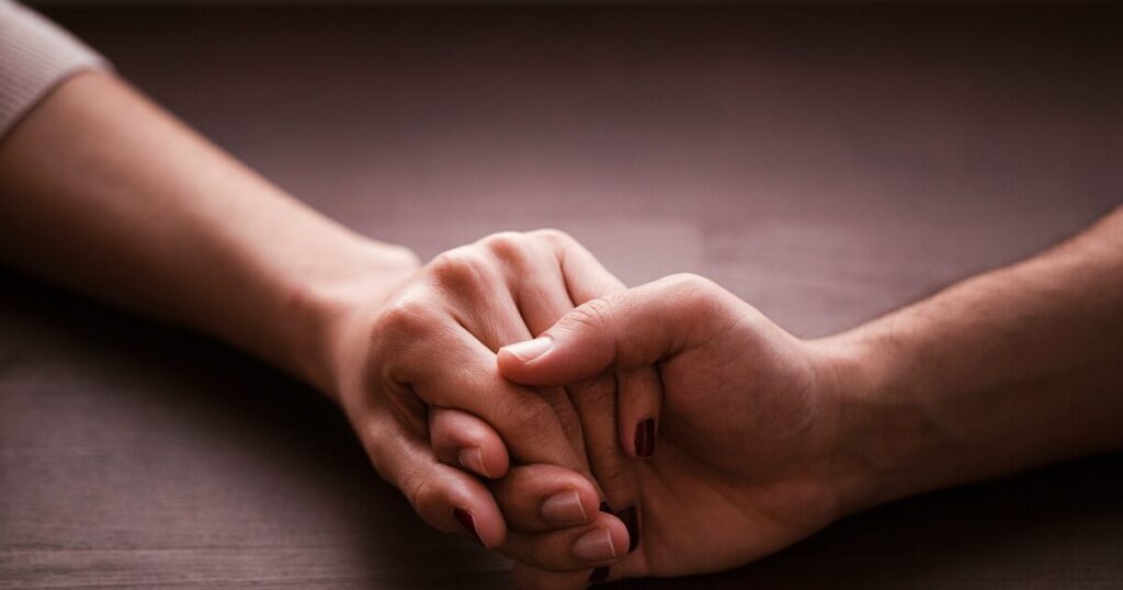 Two people hold hands in a comforting grip on a dark wooden surface.