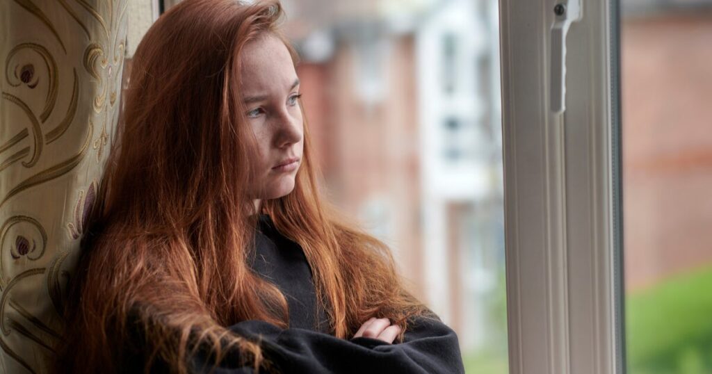 Young woman with long red hair sits by a window, gazing outside with a thoughtful expression.