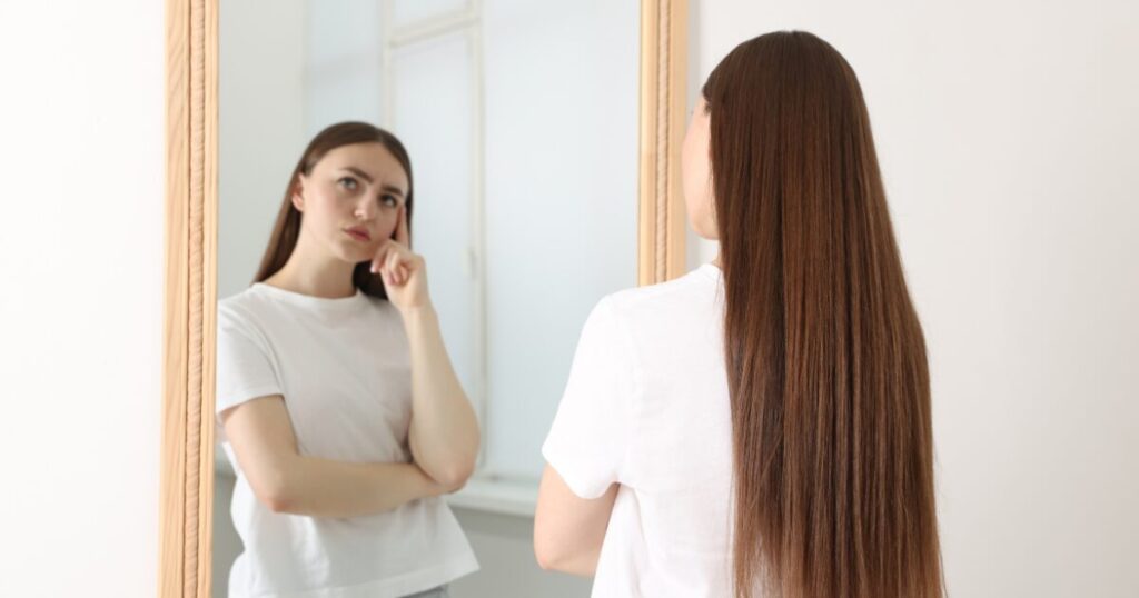 Woman with long brown hair looking at herself in a mirror, resting her chin on her hand in a white T-shirt.
