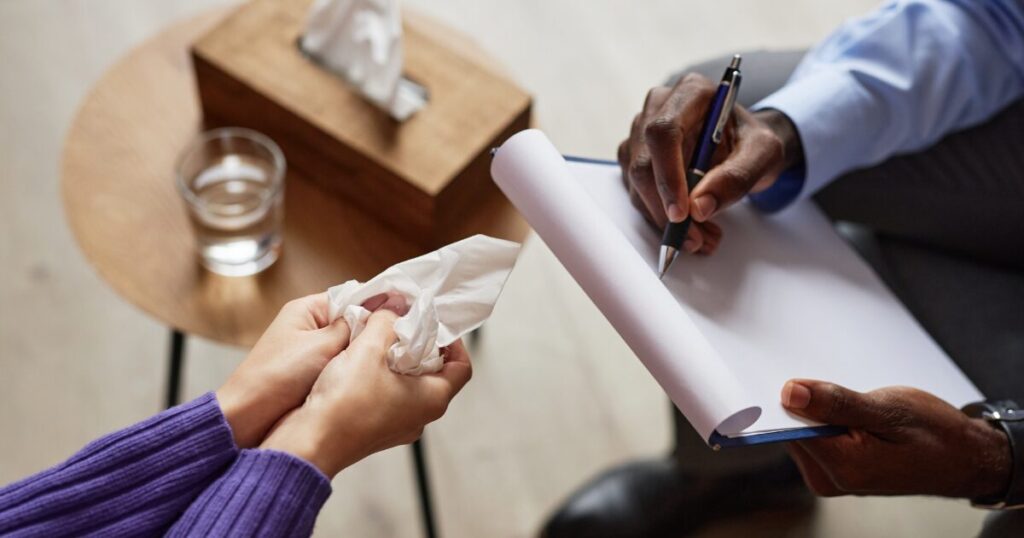 Two people in a meeting: one hands a tissue to the other while the second person writes on a clipboard with a pen nearby.