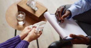 Two people in a meeting: one hands a tissue to the other while the second person writes on a clipboard with a pen nearby.