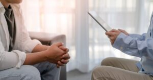 Therapist uses a tablet while a client sits with hands clasped, in a counseling setting.