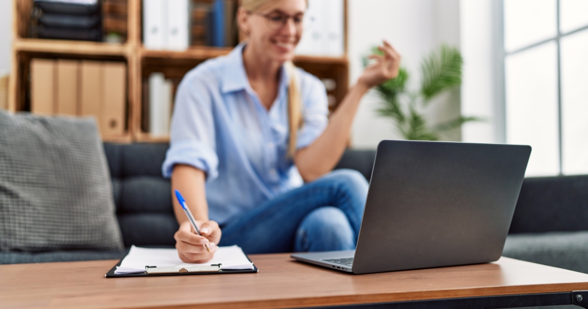 Woman sitting on a couch, taking notes on a clipboard while a laptop rests on a coffee table.