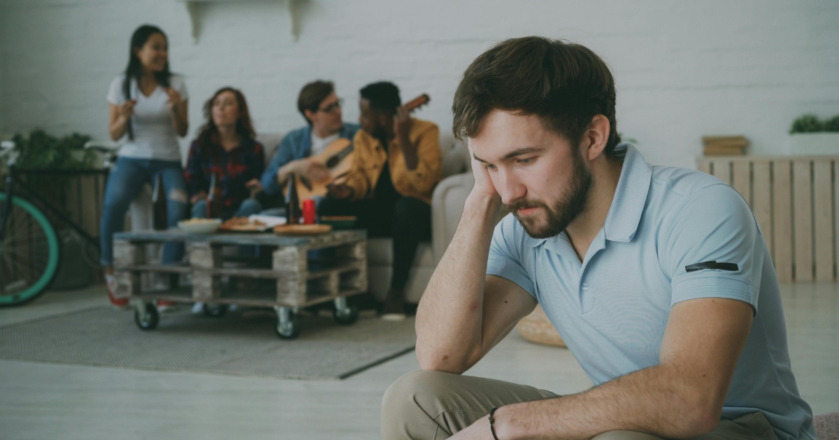 Man sitting alone in foreground, looking down while a group socializes in the background.