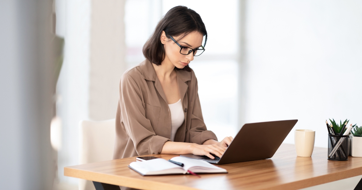 Woman with glasses typing on a laptop at a wooden desk, notebook open beside her.