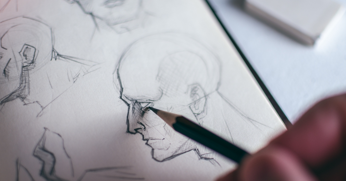 Close-up of a pencil sketch of a man's profile on paper, with a hand holding a pencil near the mouth area.