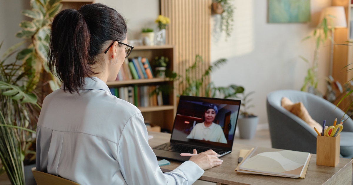 Woman with dark hair in a light blue blouse sits at a wooden desk, talking to someone on a laptop during a video call in a bright, plant-filled home office.