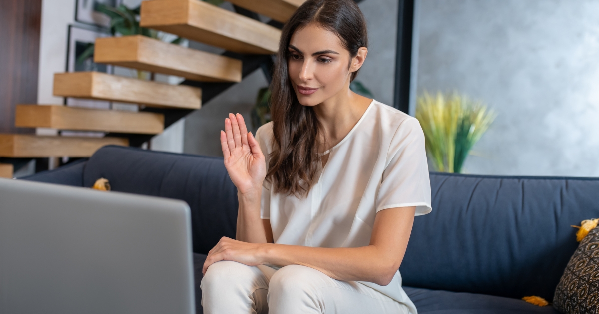 Woman seated on a blue sofa, waving hello at her laptop during a video call or online meeting.
